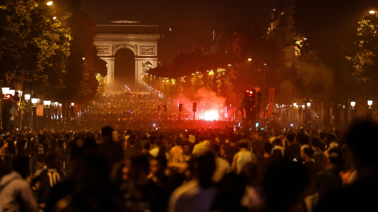 Bustling crowds swamp Avenue des Champs-Élysées, lit up by a ball of red flares along the landmark mall. The Arc de Triomphe can be seen at the end of the avenue.