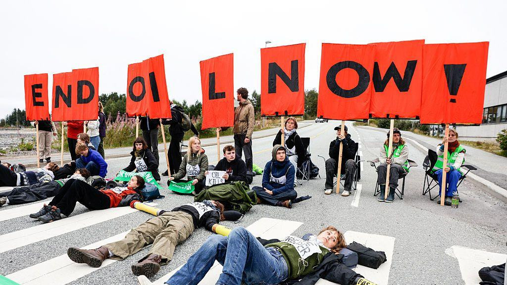 Grupa mladih ljudi protestuje protiv eksploatacije nafte u Norveškoj. Neki leže na asfaltu, dok drugi sede na stolicama i drže natpise koji zajedno formiraju poruku na engleskom: end oil now.