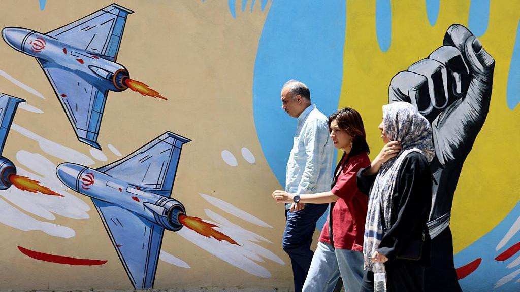 Stariji muškarac i dve žene idu ulicom pored grafita sa avionima iranske vojske