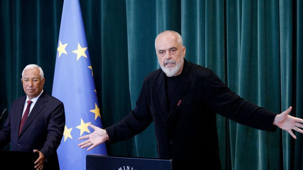 edi rama u crnoj majici i pantalonama, iza njega sa leve strane sedi muškarac u odelu i kravati