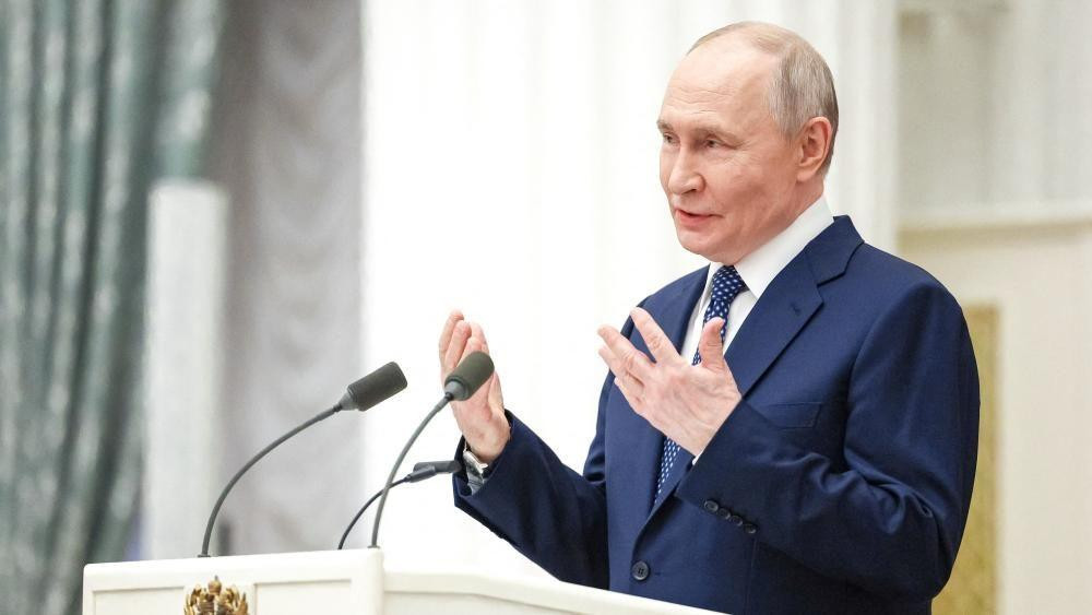 Predsednik Putin, u beloj košulji, plavoj kravati sa tufnama i plavom odelu, gestikulira rukama dok govori sa belog podijuma sa dva mikrofona. Svetlozelena zavesa sa šarom nalazi se levo od slike.