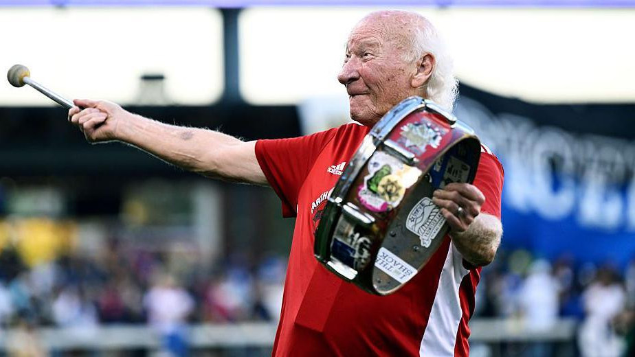 Stariji čovek sa dugom belom kosom nosi crveno-belu sportsku majicu i udara u bubanj koji drži u levoj ruci, dok se osmehuje i gleda u publiku na sportskom stadionu