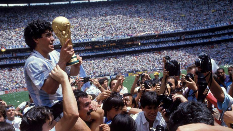 Argentinski kapiten, Dijego Maradona, drži Svetski kup u rukama i slavi osvajanje turnira 1986. godine na prepunom stadionu. Ljudi ga fotografišu. U pozadini su hiljade ljudi na tribinama i delić fudbalskog terena