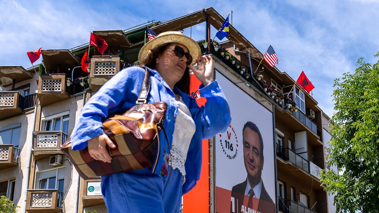 Žena ide ulicom sa naočalama za sunce iza nje plakat premijera Kosova Aljbina Kurtija i zastave Kosova, Amerike i Albanije