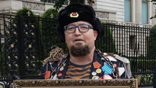 A man wearing an old-style Russian hat with a Soviet badge holds the top of a framed picture and wears a jacket crammed with medals