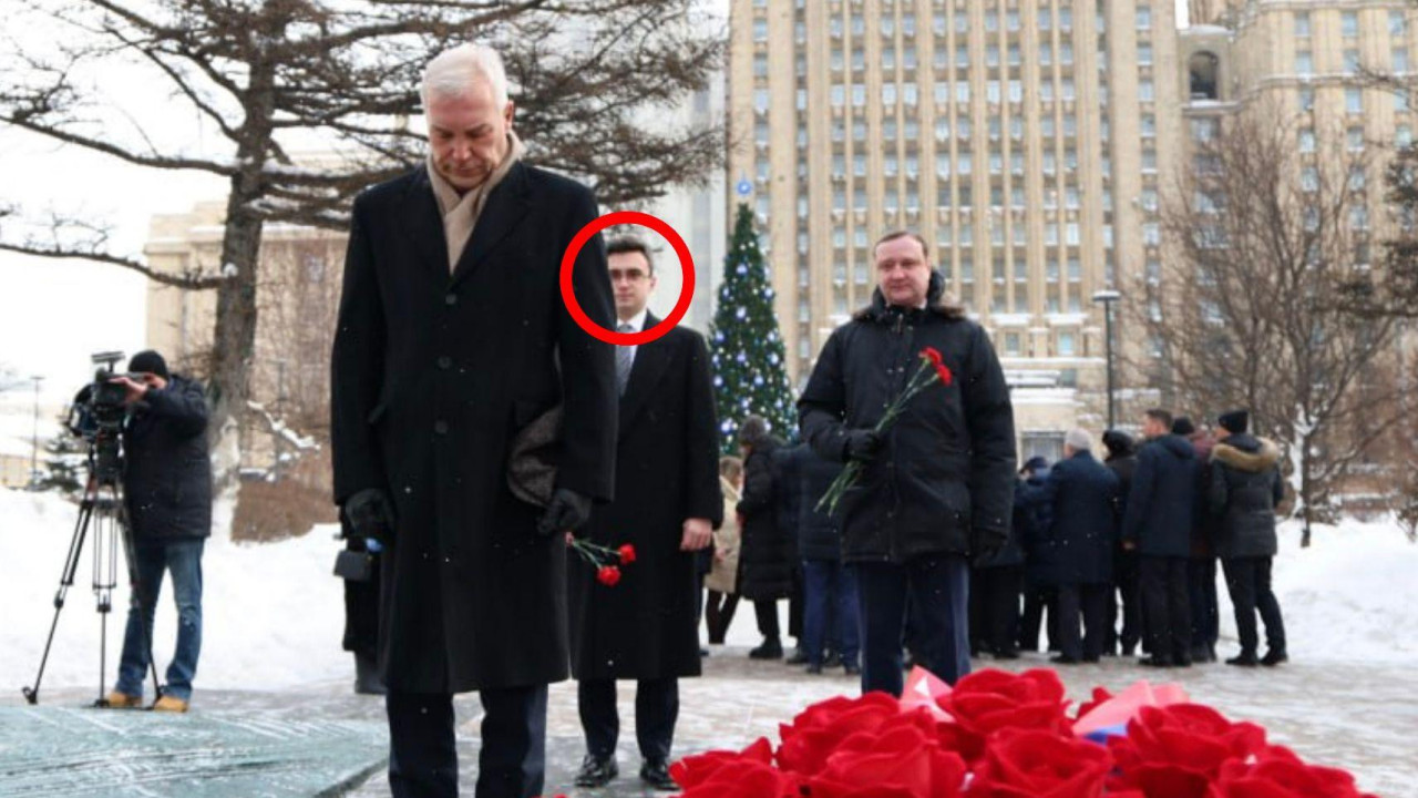 Zamenik ruskog ministra spoljnih poslova Aleksandar Gruško, čovek sa sedom kosom, u tamnom kaputu i ​​marami, stoji pognute glave ispred aranžmana crvenog cveća, naizgled položenog u znak sećanja. Iza njega stoji Jevgenij Ljukšin, mladić sa tamnom kosom i naočarima, u tamnom kaputu i ​​kravati, držeći par crvenih cvetova. Dalje iza njega, na onome što izgleda kao snežni moskovski trg, nalazi se gomila ljudi, a iza njih čovek snima događaj kamerom na stativu.