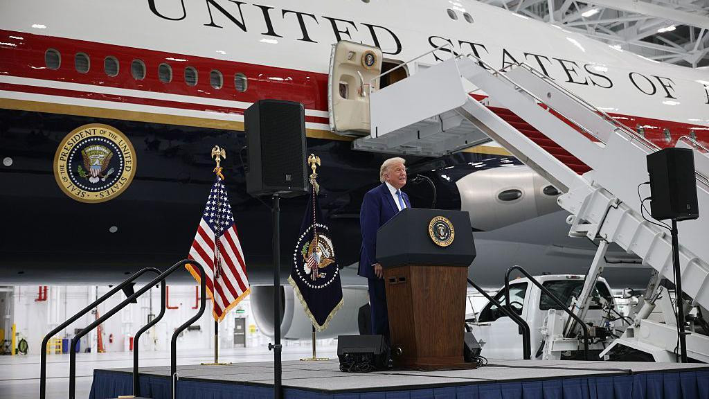 Trump standing at a podium on a stage in front of the new jet, painted white with dark red and blue stripes. There is a presidential seal and the words "United States of" visible. 