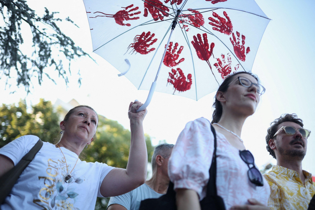 Žena drži kišobran sa crvenim otiscima ruku na njemu, ispred nje momak i devojka stoje