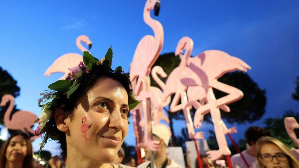 A young woman with a wreath on her head and a flamingo painted on her cheek is seen in front of flamingo-shaped placards at a protest in Albania