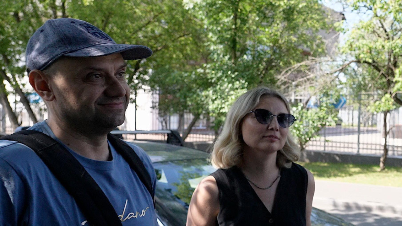 Muškarac u plavoj majici i plavim kačketom na glavi sa rancem na leđima pored kojeg je žena u crnoj haljini i naočarima za sunce