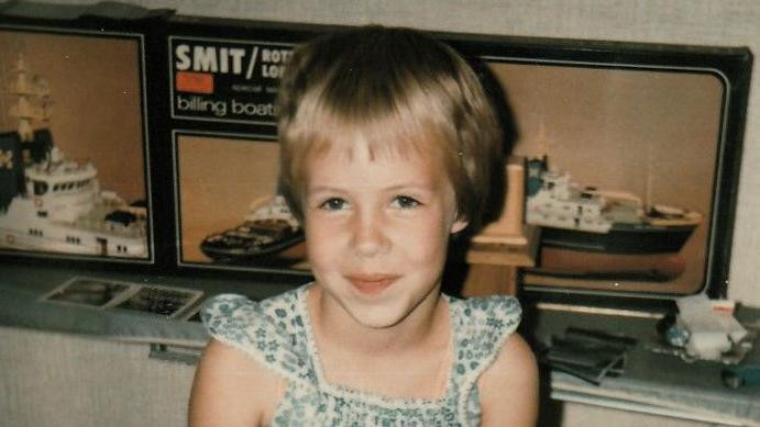 
                    A 1982 photo shows Nina Blom at seven years old, inside a house wearing a summer dress. She has short blond hair and has a shy smile on her face. She is seated in front of a display of model ships.
                