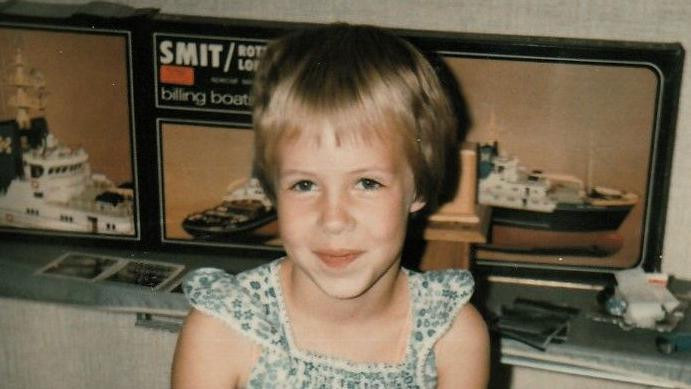 
                    A 1982 photo shows Nina Blom at seven years old, inside a house wearing a summer dress. She has short blond hair and has a shy smile on her face. She is seated in front of a display of model ships.
                