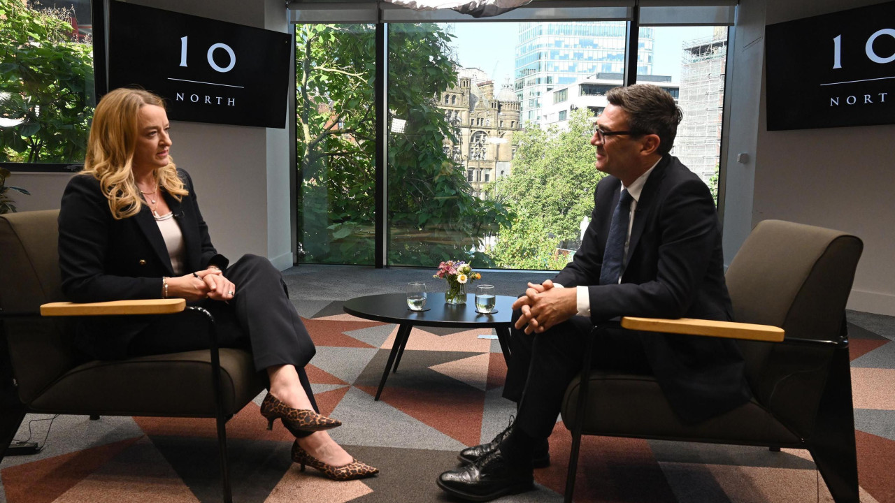 
                    Andy Burnham and Laura Kuenssberg sit opposite one another on brown chairs with a coffee table between them, on top of which are two glasses of water. There is a patterned carpet and two screens behind them which read '10 North' in large font. The back wall is entirely glass, with a view of buildin