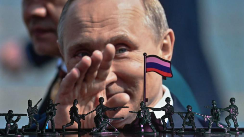 
                    U pozadini je krupni plan fotografije predsednika Putina, koji maše rukom, u prvom planu su mnoštvo tamnozelenih/crnih vojnika igračaka i ruska zastava (sa horizontalnim prugama belim (gore), plavim i crvenim (dole).
                