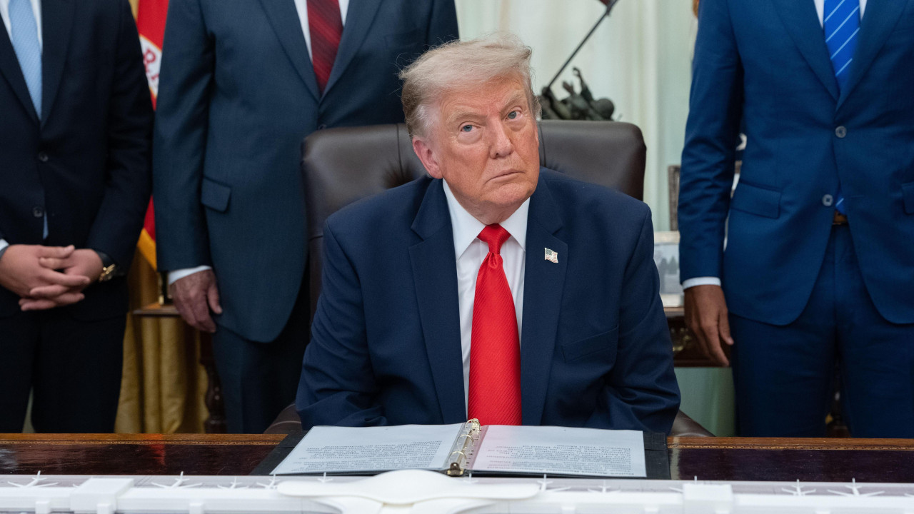 Donald Tramp u teget odelu sa crvenom kravatom u Ovalnoj sobi sedi za velikim stolom pred kojim je otvorena fascikla