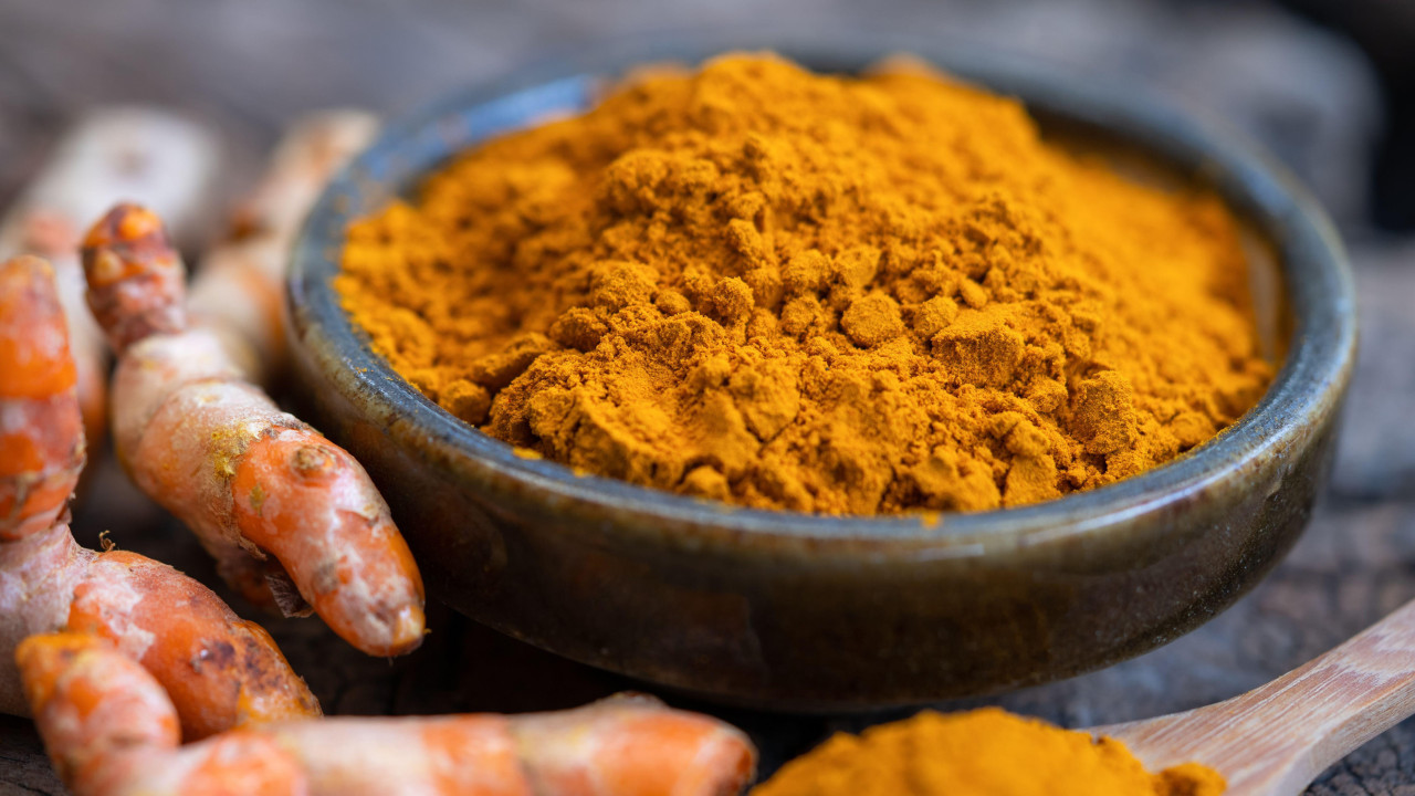 
                    Small round dish holding turmeric powder. Next to it is a wooden spoon with  small amount of powder, and to the left, small parts of the turmeric root in its raw form.
                