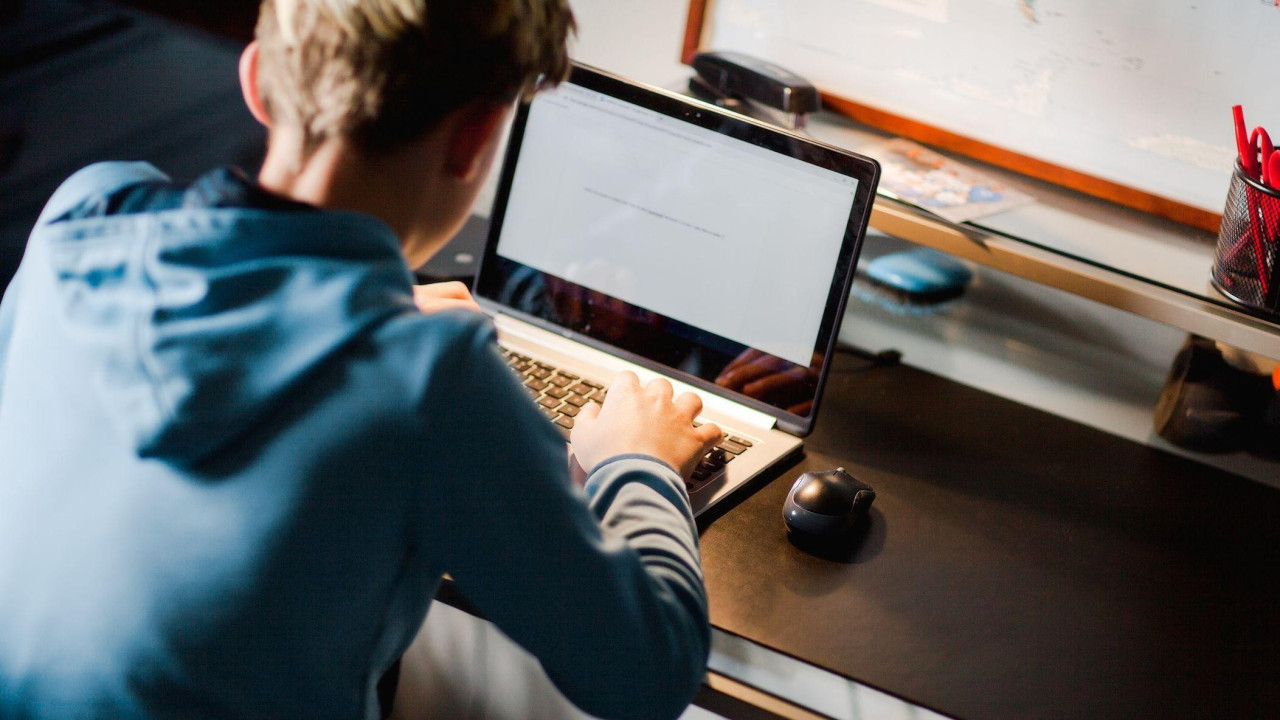 Young teenager wearing a blue hoodie pictured from behind using a laptop at a desk. 