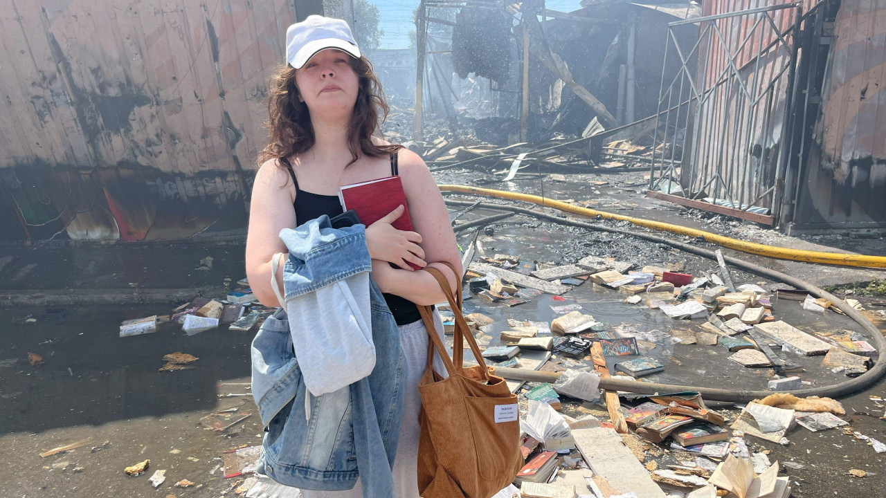 Mlada žena čvrsto drži knjigu na grudima i izgleda tužno. Iza nje je bombardovana pijačna građevina. Uništene knjige razbacane su po zemlji oko nje.