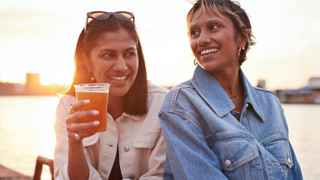 Two women are laughing together while sitting by a river with the sunset behind them; one is holding a large plastic cup of beer.