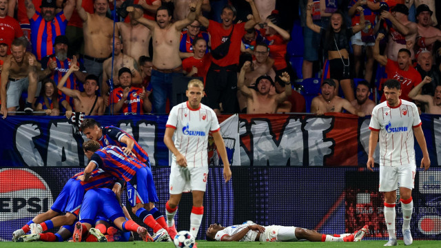 
                    fudbaleri na terenu, sa leve strane u plavo crvenim dresovima, desno dva fubalera u belim dresovima sa crvenim uspravnim linijama okrenuta ka kameri, jedan šutira loptu, između njih leži fudbaler na terenu
                