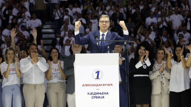 Aleksandar VUčić u plavom odelu i svetloj košulji podiže ruke u vazduh, stoji za govornicom, oko njega je mnogo ljudi