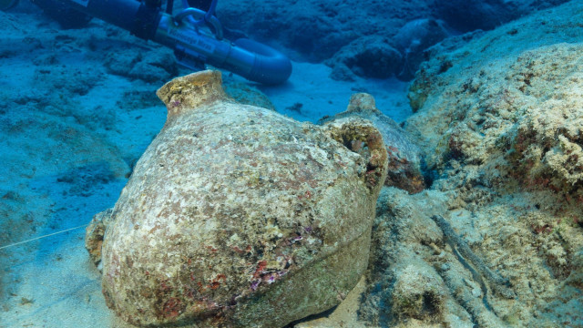 
                    A large ceramic storage pot is in the foreground and is at a slight angle leaning to the left. It has a narrow neck and two handles half way down on each side. To the right are some rocks. Behind you can just make out a diver with a large pipe that looks like a vacuum cleaner that sucks away the sa